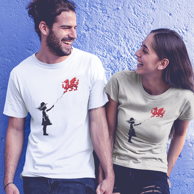 A man and a woman standing together and holding hands in front of a bright blue textured wall. The man wears a white graphic t-shirt with a red dragon balloon being held a black silhouette of a Welsh girl, paired with ripped dark jeans and white sneakers.