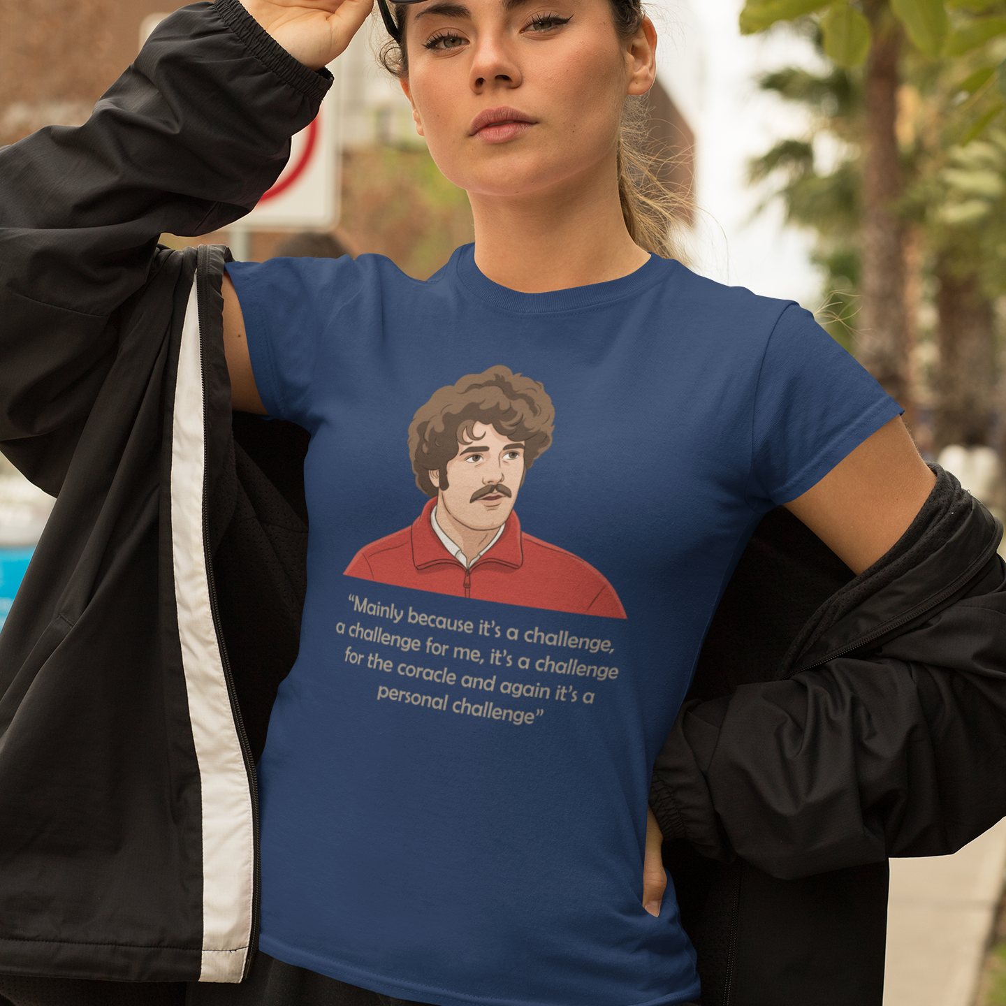 A woman wearing a blue graphic t-shirt with a printed illustration of a man and the text 'Mainly because it's a challenge, a challenge for me, it's a challenge for the coracle and again it's a personal challenge'. She is wearing a black jacket off her shoulders and sunglasses on her head, posing outdoors.