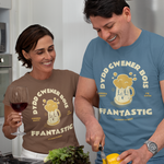 A man and a woman in a modern kitchen, both wearing matching brown and blue t-shirts with a beer mug graphic and the text 'DDYD GWENER BOIS FFANTASTIG'. The man is slicing a yellow bell pepper on a wooden cutting board, while the woman holds a glass of red wine.
