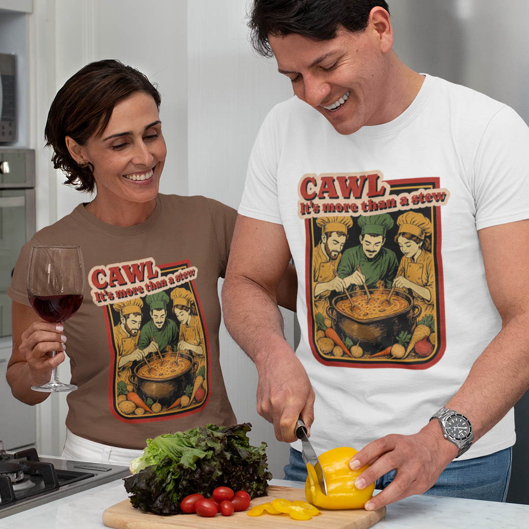 A man and a woman in a modern kitchen, both wearing matching graphic t-shirts featuring a cartoon character in a pot with the text 'CAWL It's more than a stew'. The man is cutting a yellow bell pepper on a wooden board, while the woman holds a glass of red wine.