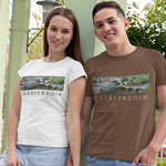 A man and a woman standing outdoors in a park-like setting with light green pillars, wearing matching graphic t-shirts. The man wears a brown t-shirt and the woman wears a white t-shirt, both featuring a landscape print with the text 'CAERFYRDDIN'.