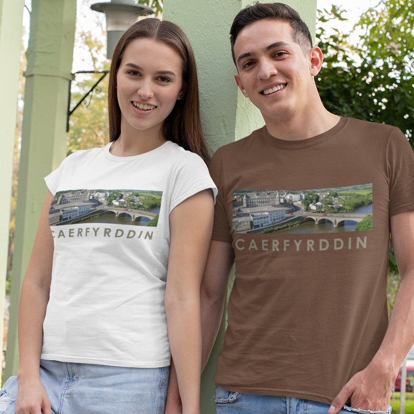 A man and a woman standing outdoors in a park-like setting with light green pillars, wearing matching graphic t-shirts. The man wears a brown t-shirt and the woman wears a white t-shirt, both featuring a landscape print with the text 'CAERFYRDDIN'.