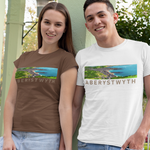 A man and a woman standing outdoors in a park-like setting with light green pillars, wearing matching graphic t-shirts. The woman's shirt is brown with a landscape print, and the man's shirt is white with a blue landscape print and the text 'ABERYSTWYTH'.