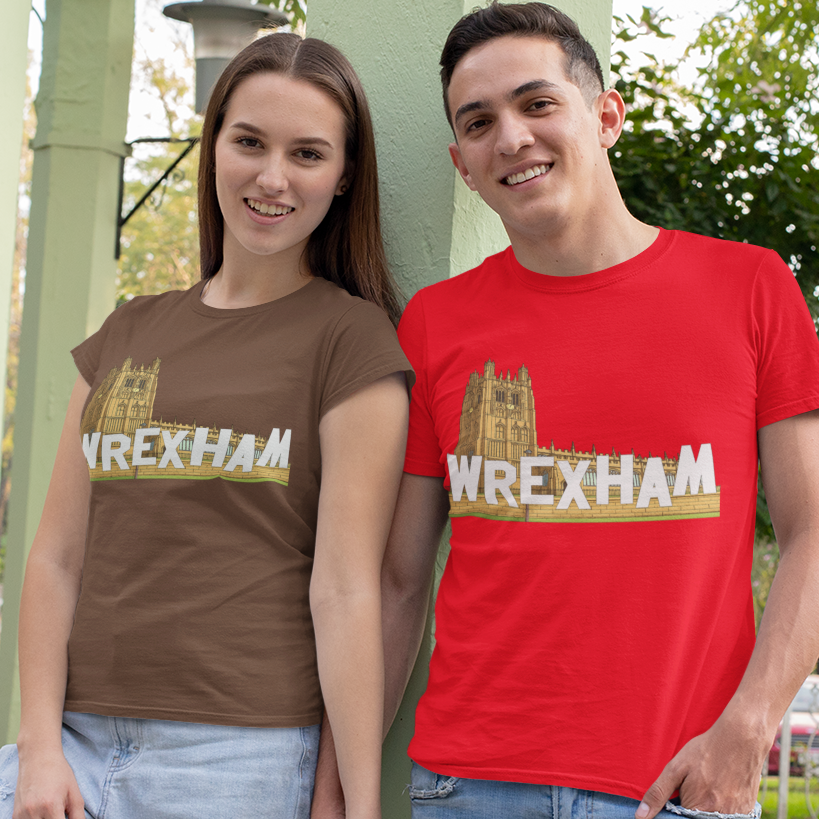 A man and a woman standing outdoors in a park-like setting, both wearing matching graphic t-shirts with 'WREXHAM' printed across the chest. The woman's shirt is brown, and the man's shirt is bright red.