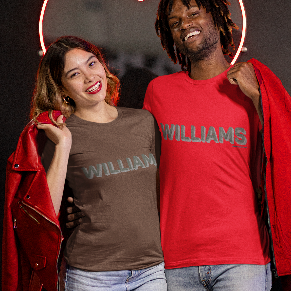 Two models posing in matching graphic t-shirts with 'WILLIAMS' printed across the chest, one in brown and one in red, each holding a red jacket, set against a dark background with a glowing heart-shaped neon sign.