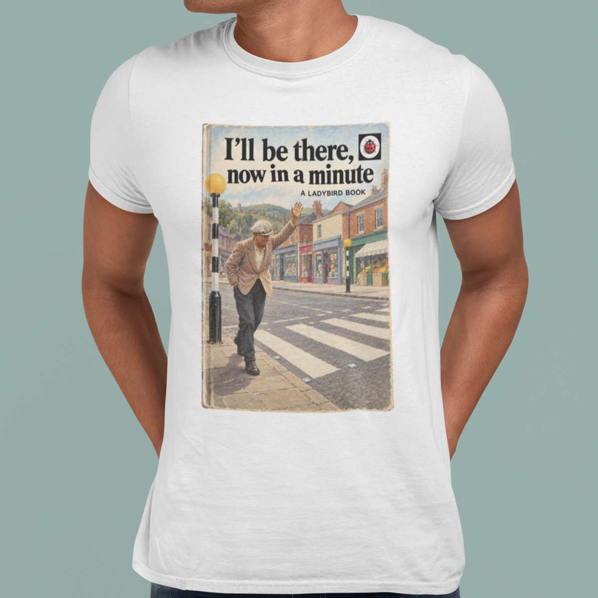 A man wearing a white crewneck t-shirt with a vintage-style London street crosswalk advertisement graphic printed on the chest, paired with dark blue jeans, set against a solid muted green background.