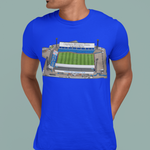 A man wearing a bright blue t-shirt with a graphic print of a football stadium on the front, paired with dark blue jeans, set against a solid muted green background.