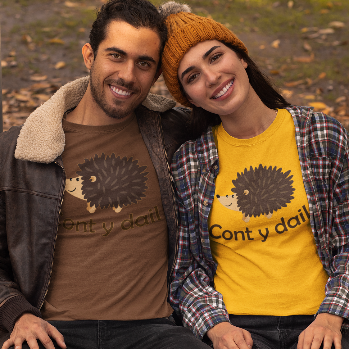 A man and a woman sitting together on a park bench, both wearing matching graphic t-shirts with a hedgehog design and the text 'Cont y dail'. The man wears a brown leather jacket over a brown t-shirt, and the woman wears a yellow t-shirt under a red plaid flannel shirt and a mustard beanie.