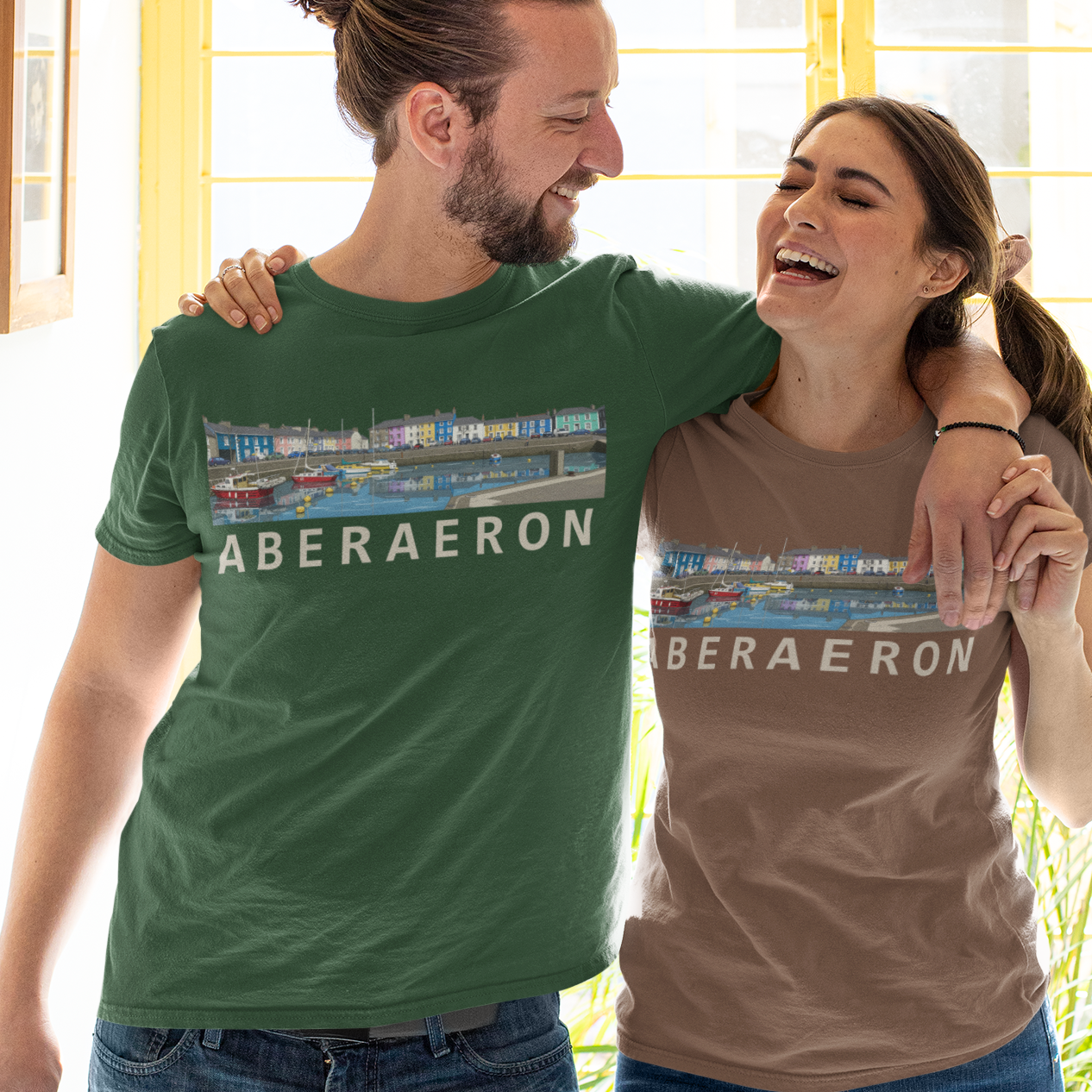 A man and a woman standing together indoors, both wearing matching graphic T-shirts with the word 'ABERAERON' printed across the chest. The man's shirt is green and the woman's shirt is taupe, and both feature a colorful rectangular graphic above the text.
