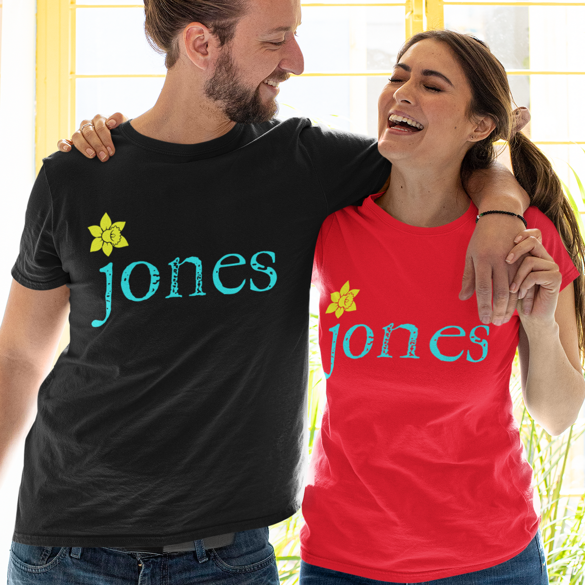 A man and a woman standing together indoors, each wearing a custom graphic t-shirt. The man wears a black t-shirt with 'jones' in teal and a yellow daffodil, while the woman wears a red t-shirt with the same design.