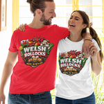 A man and a woman standing arm in arm, both wearing matching graphic T-shirts with a 'Welsh Bollocks' logo. The man wears a red T-shirt and the woman wears a white T-shirt, both paired with blue jeans in front of a bright yellow window.