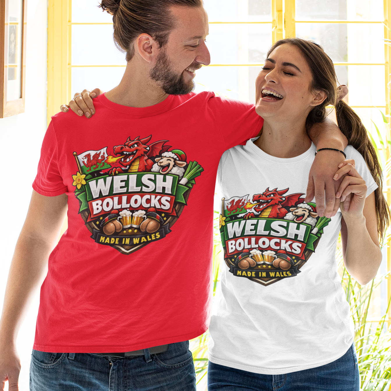 A man and a woman standing arm in arm, both wearing matching graphic T-shirts with a 'Welsh Bollocks' logo. The man wears a red T-shirt and the woman wears a white T-shirt, both paired with blue jeans in front of a bright yellow window.