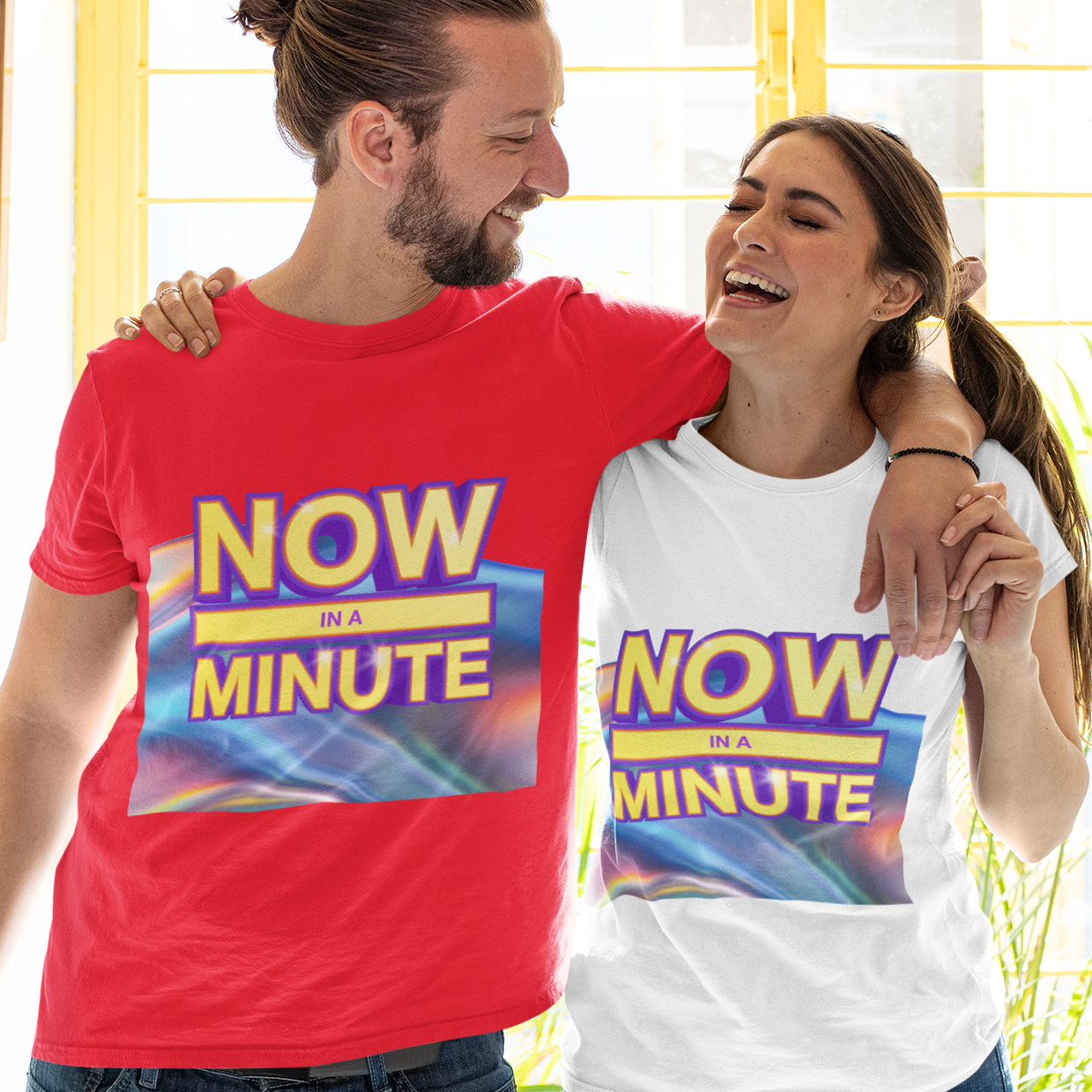 A man and a woman standing together indoors, both wearing matching graphic T-shirts with the slogan 'NOW IN A MINUTE' in yellow and blue. The man's shirt is bright red, and the woman's shirt is white.
