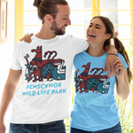 A man and a woman standing together in front of a bright yellow window, wearing matching graphic t-shirts. The man wears a white t-shirt and the woman wears a light blue t-shirt, both featuring a colorful wild life illustration and the text 'PENSCYNOR WILDLIFE PARK'.