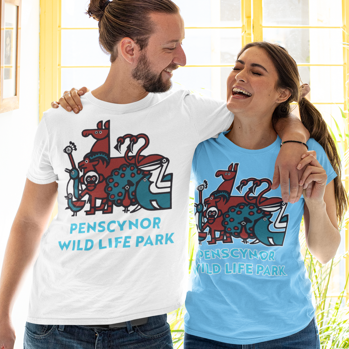A man and a woman standing together in front of a bright yellow window, wearing matching graphic t-shirts. The man wears a white t-shirt and the woman wears a light blue t-shirt, both featuring a colorful wild life illustration and the text 'PENSCYNOR WILDLIFE PARK'.