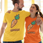 A man and a woman standing arm-in-arm in front of a yellow window, wearing matching graphic t-shirts. The man's shirt is bright yellow and the woman's shirt is bright orange, both featuring a cartoon carrot graphic and the word 'MORON' printed below it.
