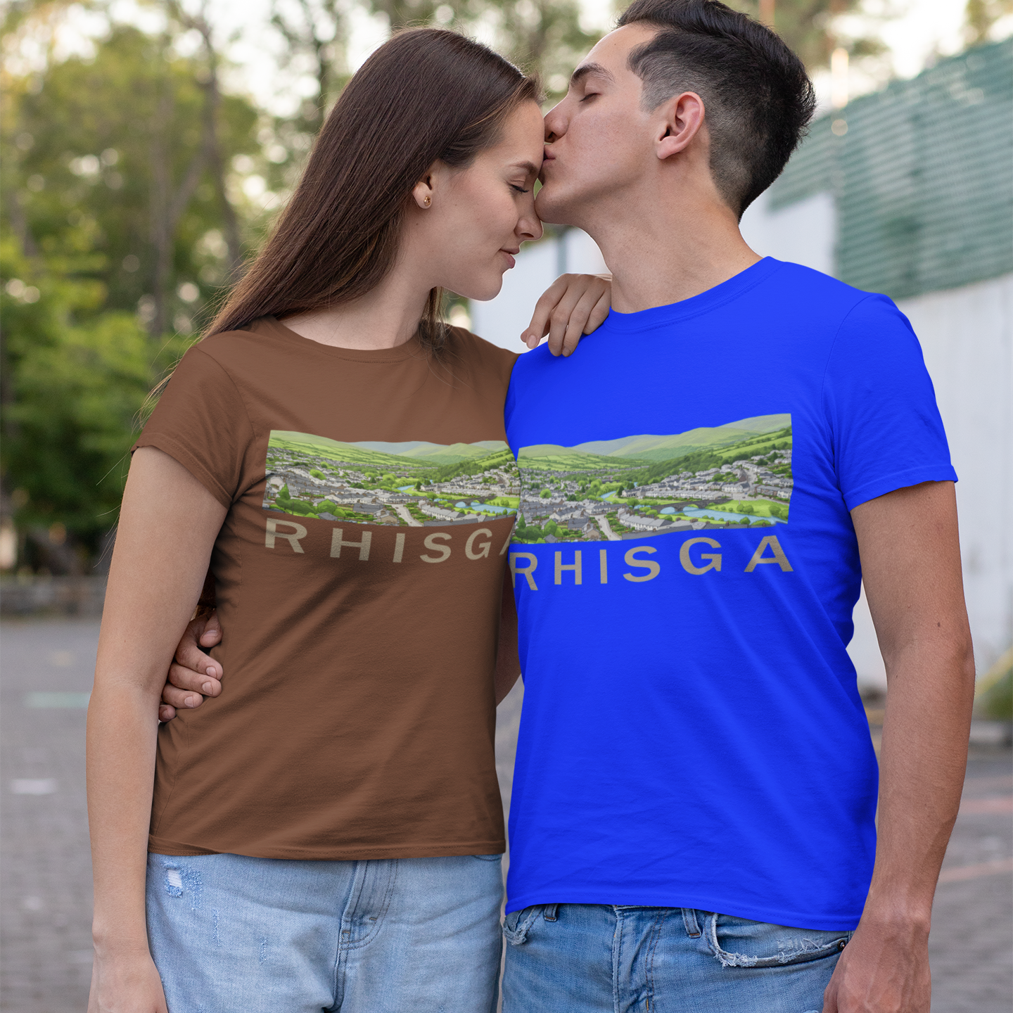 A couple outdoors wearing matching graphic t-shirts. The woman wears a brown t-shirt and the man wears a bright blue t-shirt, both featuring a landscape graphic and the text 'RHISGA'.