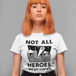 A model with orange hair wearing a white graphic t-shirt that reads 'NOT ALL HEROES WEAR CAPES' with a black and gray illustration, paired with black jeans, against a solid gray background.
