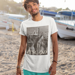 A man standing on a sandy beach wearing a white graphic t-shirt with a black and white photo print and the text 'PYSG-OD', paired with turquoise swim shorts. Boats are visible in the background under bright sunlight.