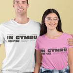 A man and a woman modeling matching graphic t-shirts against a solid pale yellow background. The man wears a white t-shirt and the woman wears a pink t-shirt, both featuring the black text 'IN CYMRU I FIND PEACE' with Welsh flag elements.