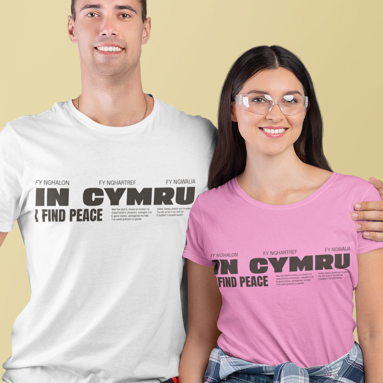 A man and a woman modeling matching graphic t-shirts against a solid pale yellow background. The man wears a white t-shirt and the woman wears a pink t-shirt, both featuring the black text 'IN CYMRU I FIND PEACE' with Welsh flag elements.
