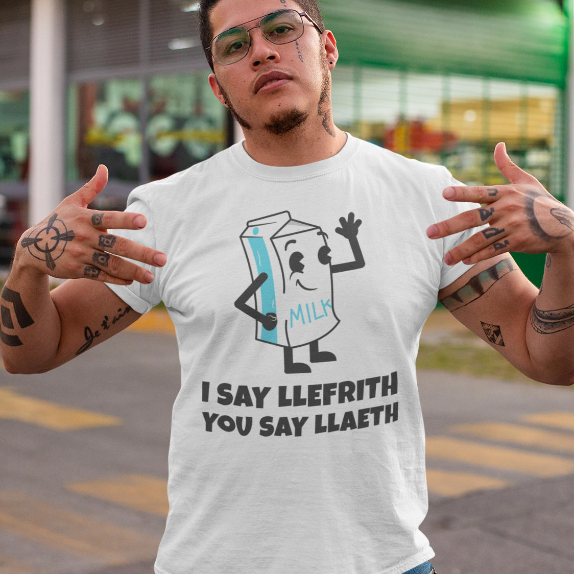 A man wearing a white graphic t-shirt with a cartoon milk carton character and the text 'I SAY LLEFRITH YOU SAY LLAETH' stands outdoors in front of a store.