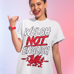 A woman modeling a white graphic t-shirt with the text 'WELSH NOT ENGLISH' and a red dragon illustration, set against a pink and purple gradient background.