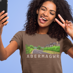 A woman with curly dark hair takes a selfie while wearing a brown graphic t-shirt that reads 'ABERMAGWR' with a road scene print. She is posing with peace signs against a solid light blue background.