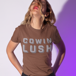 A woman wearing a brown graphic t-shirt with 'COWIN LUSH' printed in gray block letters, paired with black jeans, posing against a white wall with a purple shadow cast on the right.