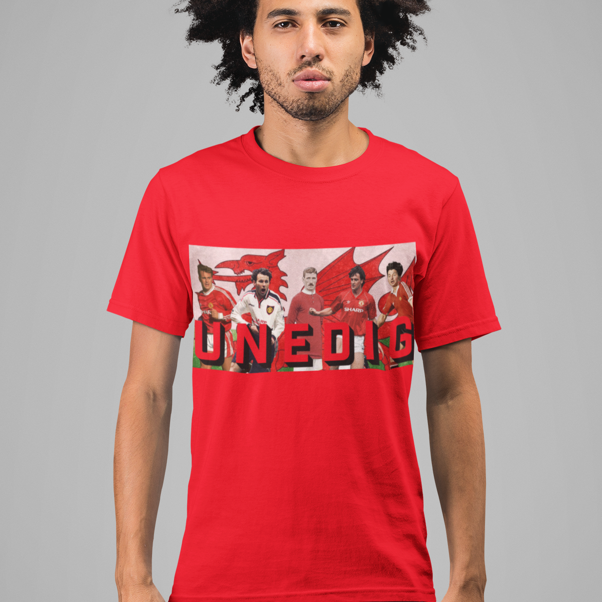 A man with dark curly hair wearing a bright red graphic t-shirt and dark blue jeans, standing against a solid gray studio background. The t-shirt features a rectangular collage graphic of football players with the word 'UNEDIG' printed in bold red letters.