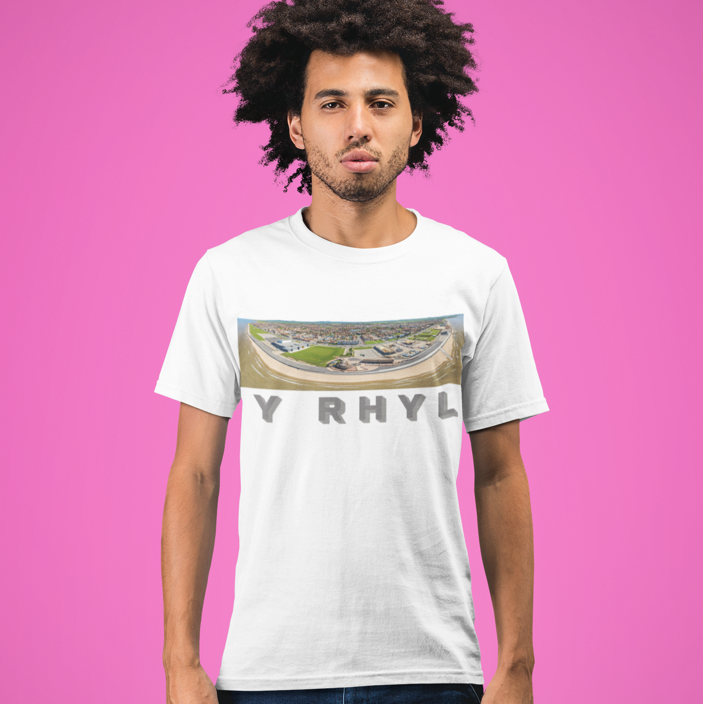 A man with dark curly hair wearing a white graphic t-shirt and dark blue jeans, standing against a solid pink background. The t-shirt features a colorful stadium graphic above the text 'Y RHYL'.