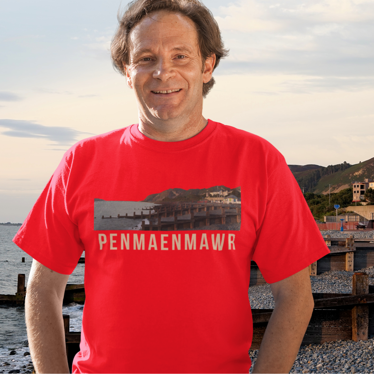 Man wearing a red t-shirt with 'Penmaenmawr' printed on it, standing by a coastal area.