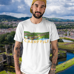 A man wearing a white graphic t-shirt with a 'CAERFFILI' print, khaki pants, and a gold cap, set against a scenic background of Caerphilly castle and river.