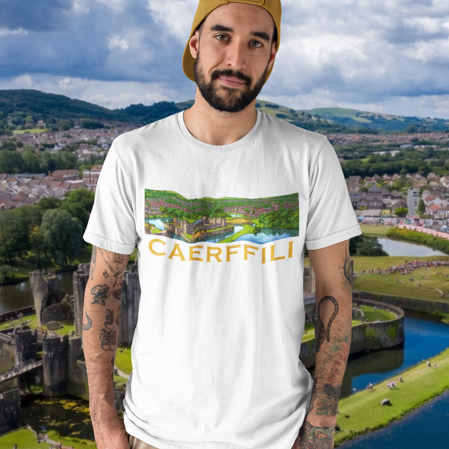 A man wearing a white graphic t-shirt with a 'CAERFFILI' print, khaki pants, and a gold cap, set against a scenic background of Caerphilly castle and river.