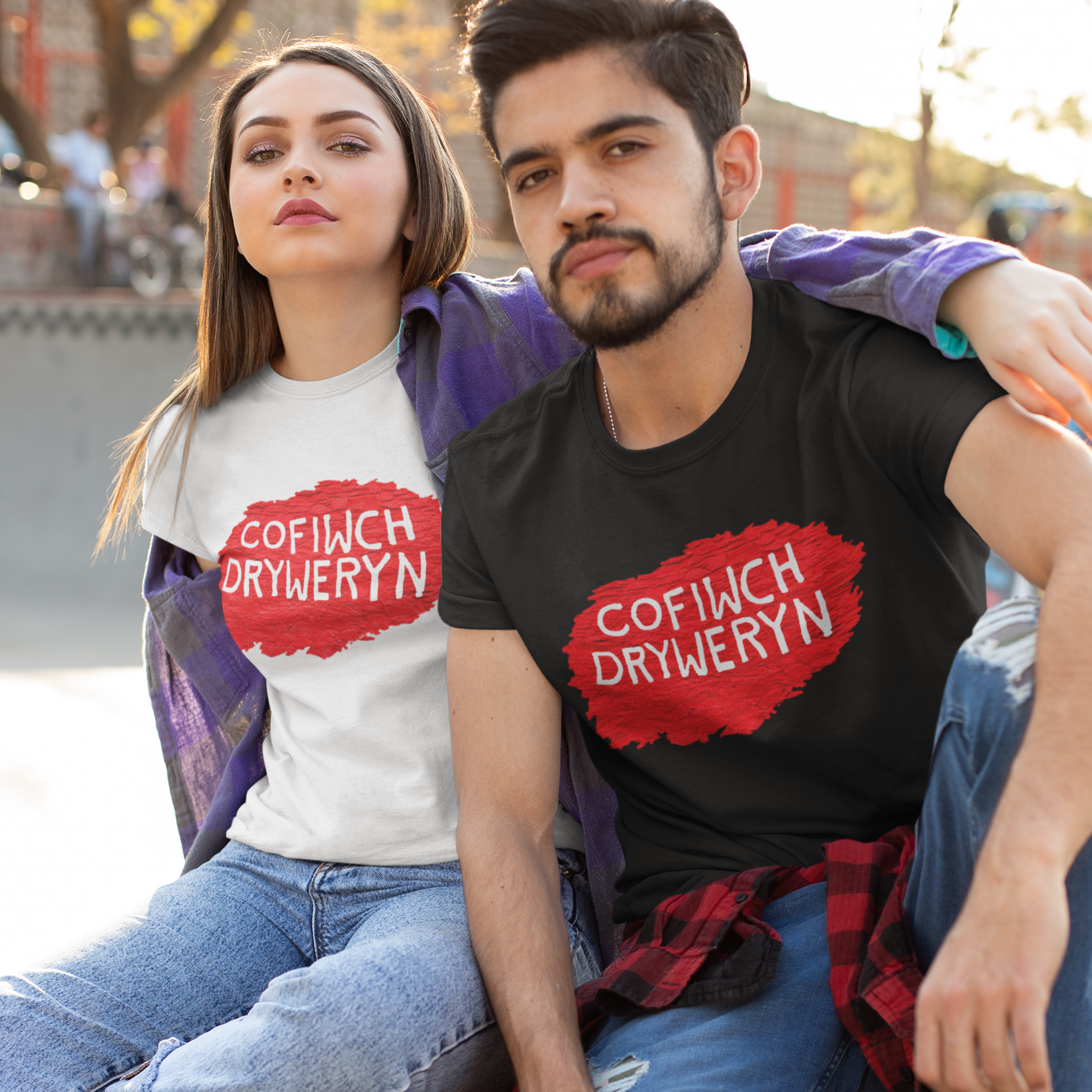 Two models sitting together at a skatepark wearing matching graphic t-shirts. The model on the left wears a white t-shirt with a red paint-splatter logo containing the Welsh text 'COFIWCH DRYWYREN', and the model on the right wears a black t-shirt with the same red logo.