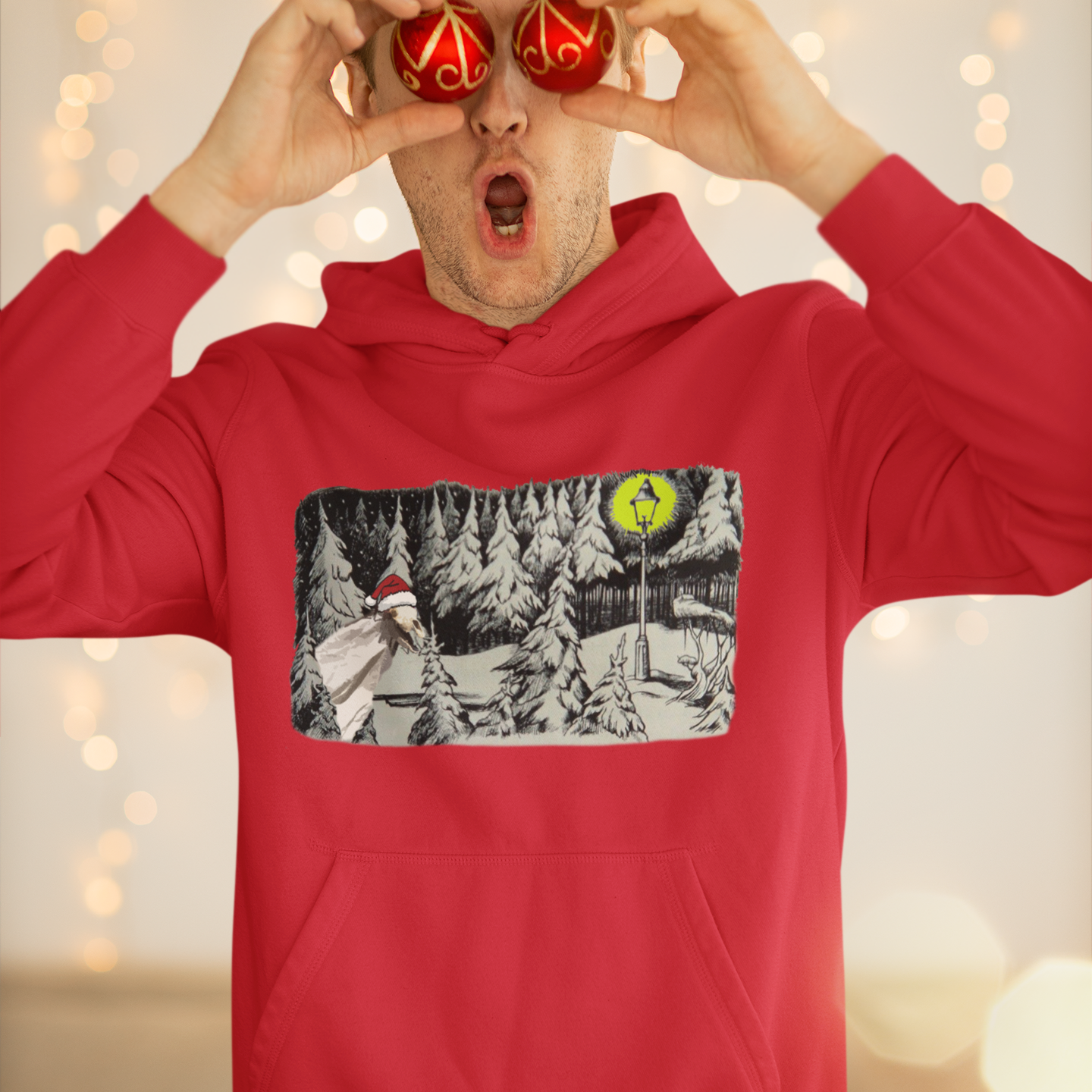 Person wearing a red hoodie with a graphic design, holding small red ornaments to their eyes against a blurred light background.